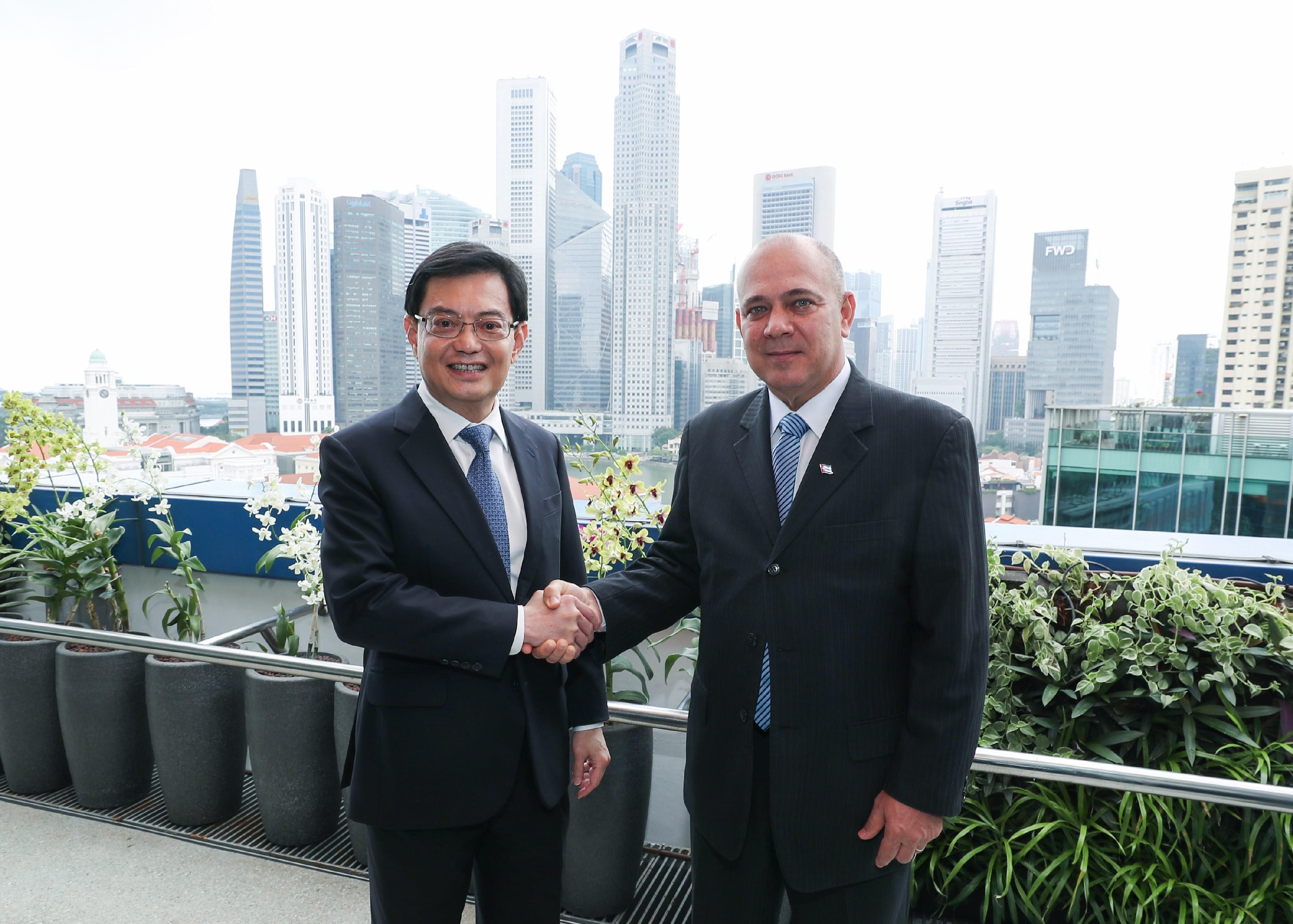 Two men in suits shake hands on a balcony, skyscrapers in the background.